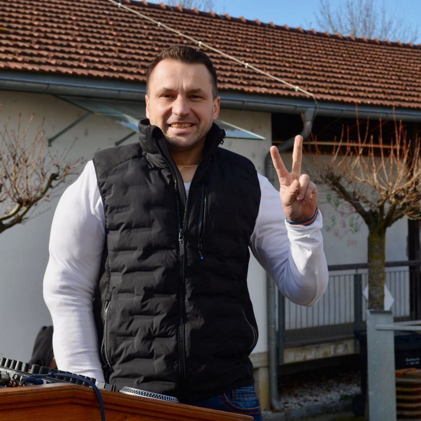 Ein Mann steht lächelnd vor einem Haus mit Ziegeldach und macht ein Friedenszeichen. Er trägt eine schwarze Weste über einem weißen Hemd.