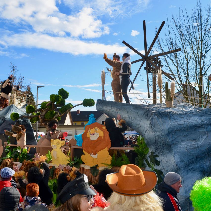 Ein Paradewagen mit Menschen in Kostümen, einschließlich eines Löwen, einer Windmühle und eines Baumes, zieht eine Menge an. Es ist vor einem blauen Himmel mit Wolken.