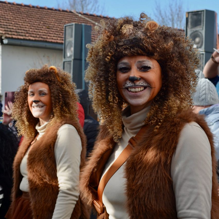 Zwei Frauen in Löwenkostümen stehen zusammen und lächeln für ein Foto im Freien. Hinter ihnen befindet sich ein Gebäude mit einem Ziegeldach, einem Lautsprecher und anderen Menschen.