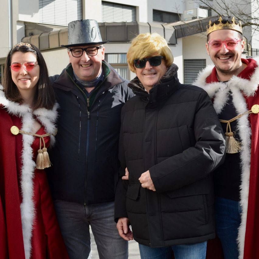 Vier Personen stehen vor einem Gebäude. Drei tragen rote Umhänge und Kronen. Eine Person hat einen schwarzen Hut und eine Brille.