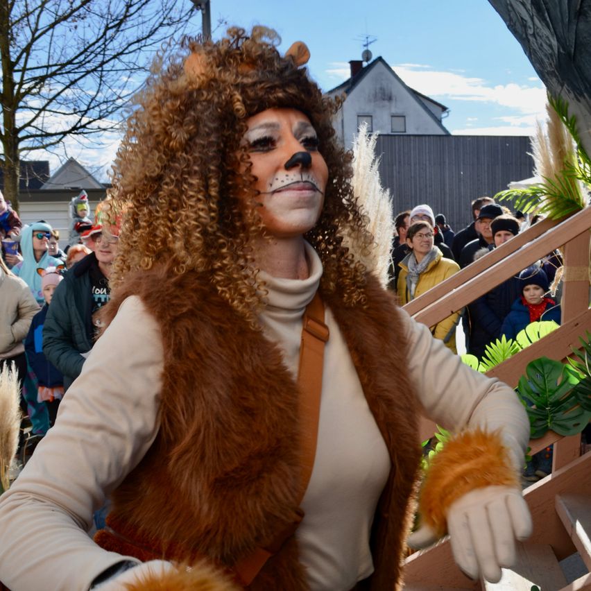 Eine Person in Löwenkostüm steht an einem sonnigen Tag vor einer Menge. Ein Haus mit Schornstein ist hinter der Menge sichtbar.