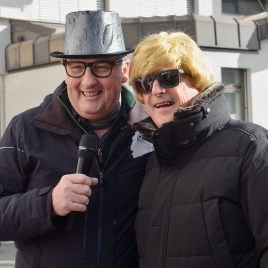 Zwei Männer stehen nebeneinander im Freien, einer hält ein Mikrofon. Sie tragen beide dunkle Mäntel. Der Mann auf der linken Seite hat einen silbernen Zylinderhut und eine Brille. Der Mann auf der rechten Seite hat eine blonde Perücke und eine Sonnenbrille.