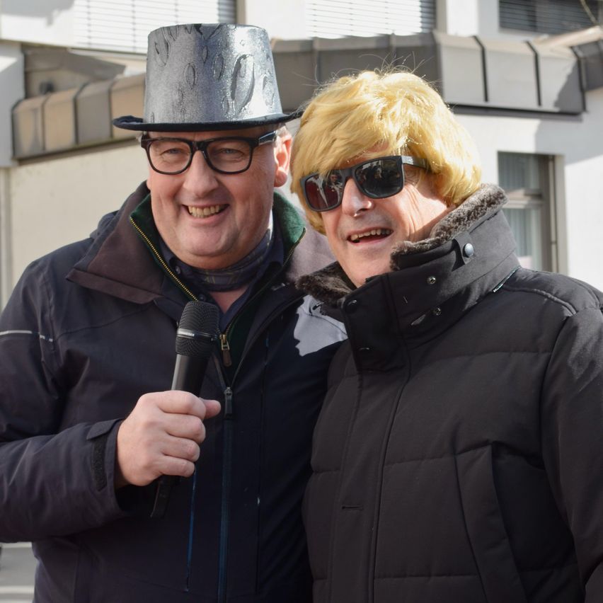 Zwei Männer stehen nebeneinander im Freien, einer hält ein Mikrofon. Sie tragen beide dunkle Mäntel. Der Mann auf der linken Seite hat einen silbernen Zylinderhut und eine Brille. Der Mann auf der rechten Seite hat eine blonde Perücke und eine Sonnenbrille.