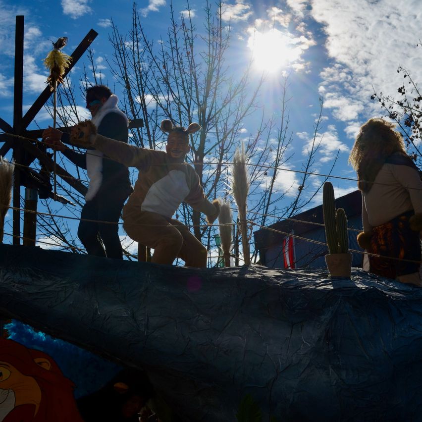 Eine Gruppe von Menschen in Kostümen steht auf einem Wagen bei einer Parade. Eine Person ist als Bär verkleidet, eine andere in einem Löwenkostüm. Der Wagen ist mit Bäumen und einem Kaktus dekoriert. Der Himmel ist blau mit einigen Wolken.