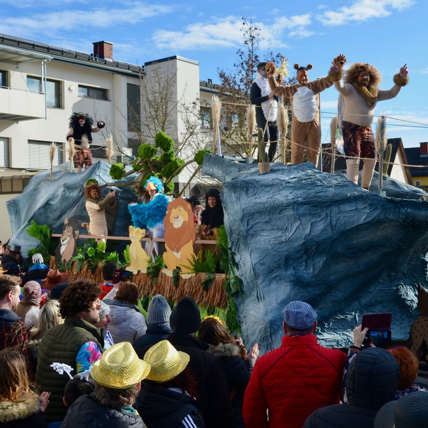 Ein Karnevalszug mit einer Felsformation, bei dem Menschen in Tierkostümen zu sehen sind, darunter Löwen und ein Affe. Zuschauer beobachten den Zug in einem öffentlichen Bereich.