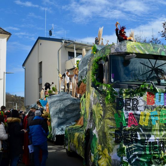 Ein Karnevalswagen, der mit Pflanzen und Tieren verziert ist, einschließlich eines Fahrers in einem roten Hut, wird vor einer Menschenmenge und Gebäuden ausgestellt.