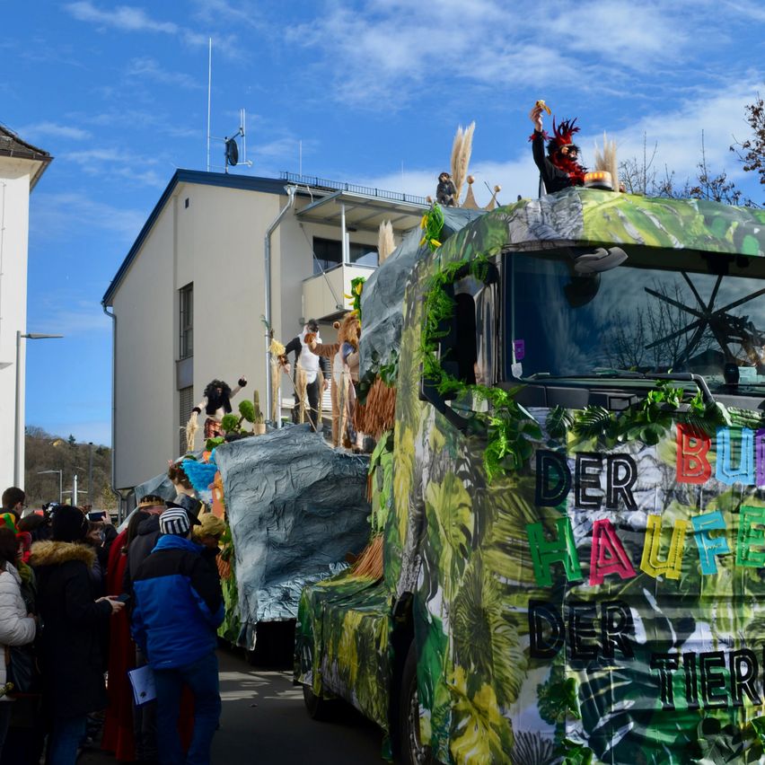 Ein Karnevalswagen, der mit Pflanzen und Tieren verziert ist, einschließlich eines Fahrers in einem roten Hut, wird vor einer Menschenmenge und Gebäuden ausgestellt.