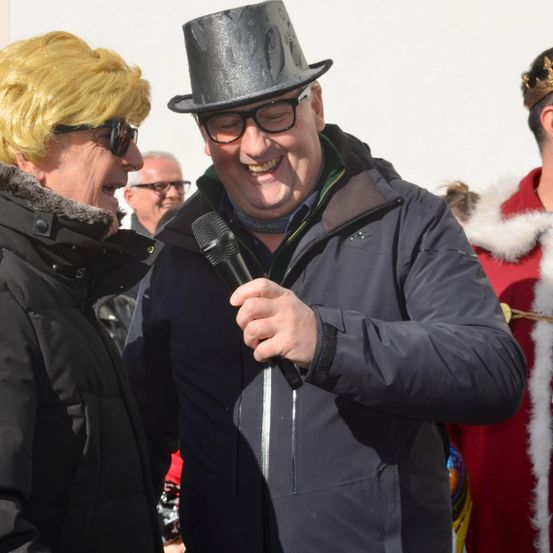 Ein Mann mit einem Zylinder und Brille hält ein Mikrofon und lächelt. Eine Frau mit blonden Haaren und einer Brille steht neben ihm. Dahinter steht ein Mann in einem roten Kostüm mit Krone.