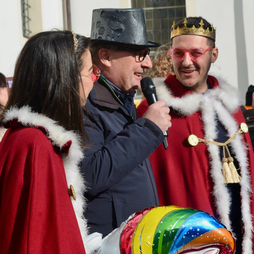 Drei Personen in festlichen Kostümen stehen draußen. Ein Mann in einem schwarzen Hut hält ein Mikrofon. Ein Mann in einem roten Gewand trägt eine goldene Krone. Eine Frau in einem roten Gewand blickt den Mann an. Ein bunter Ballon ist im Vordergrund.