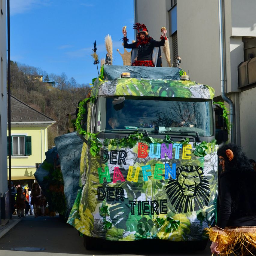 Ein dekorierter LKW mit einem Löwengrafik auf der Seite, einer Person mit Kopfschmuck obenauf und einer weiteren Person in einer Affenverkleidung, die daneben geht.