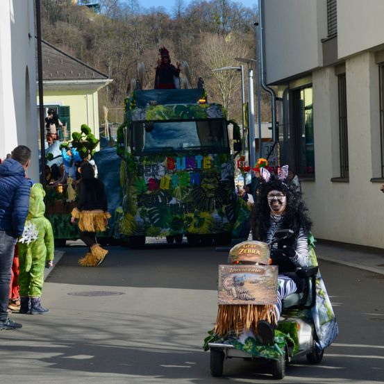 Ein festlicher Umzug mit einem dekorierten Lastwagen, auf dem Menschen in Kostümen stehen. Eine Person, die als Zebra verkleidet ist, fährt einen Golfwagen. Zuschauer in Kostümen beobachten von der Seite.