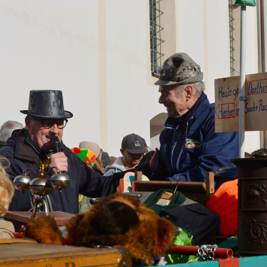 Zwei ältere Männer stehen bei einer Parade. Einer trägt einen Zylinder und der andere eine Kappe. Sie lächeln beide und einer hält ein Mikrofon. Dahinter steht eine Menschenmenge und ein Holzschild.