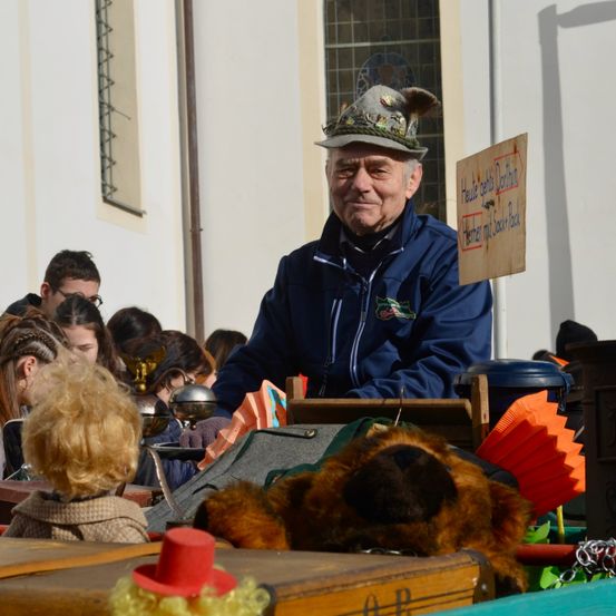 Ein älterer Mann mit einer traditionellen Mütze sitzt auf einem Wagen mit einem Kuscheltier, umgeben von Kindern. Ein Schild mit deutschem Text ist an einem Pfosten befestigt.