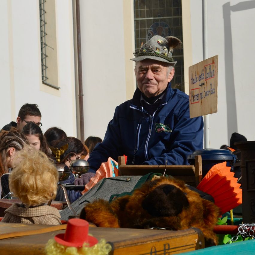 Ein älterer Mann mit einer traditionellen Mütze sitzt auf einem Wagen mit einem Kuscheltier, umgeben von Kindern. Ein Schild mit deutschem Text ist an einem Pfosten befestigt.