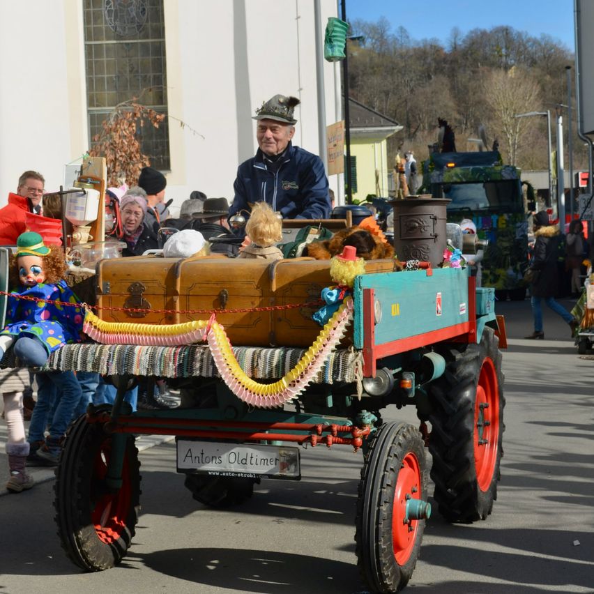 Ein älterer Mann sitzt in einem dekorierten Traktor bei einer Parade, mit Kindern und verschiedenen Gegenständen um ihn herum. Hinter ihm ist ein grüner LKW geparkt, und ein Gebäude mit Glasfenstern ist sichtbar.