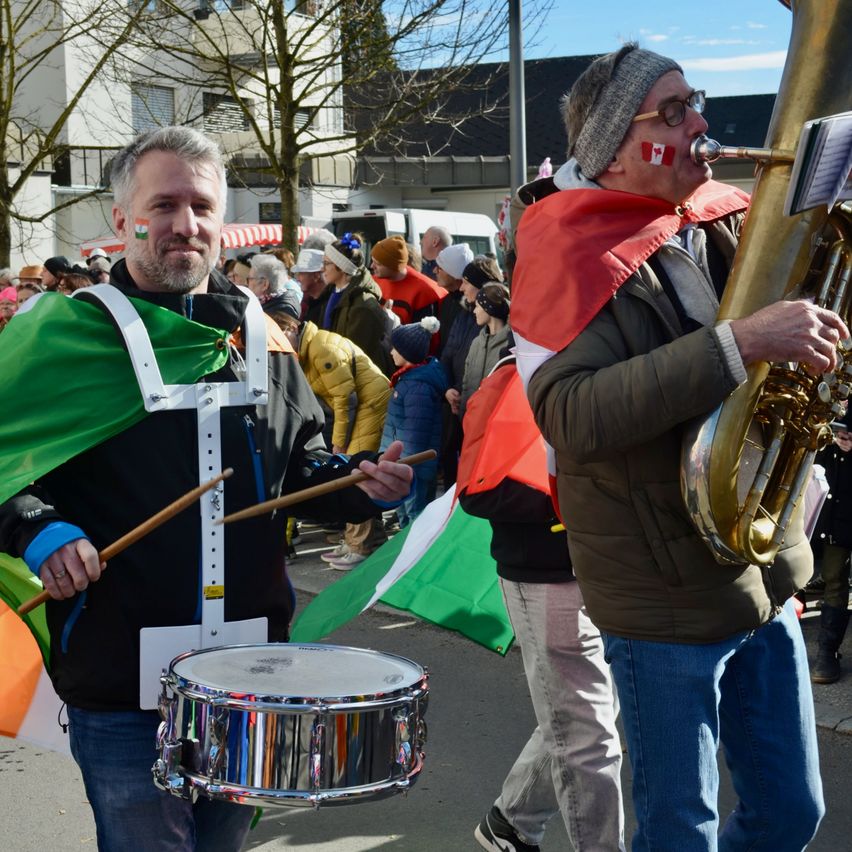 Zwei Männer marschieren in einem Umzug. Einer spielt Tuba, trägt einen irischen Flaggenumhang, und der andere spielt Trommel. Zuschauer und Gebäude sind im Hintergrund.