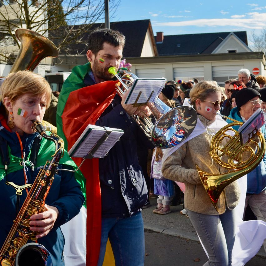 Eine Gruppe von Musikern spielt im Freien Instrumente. Einer spielt eine Saxophon, ein anderer eine Trompete und ein anderer ein Waldhorn. Sie sind vor einer Menschenmenge, einige in Kostümen.