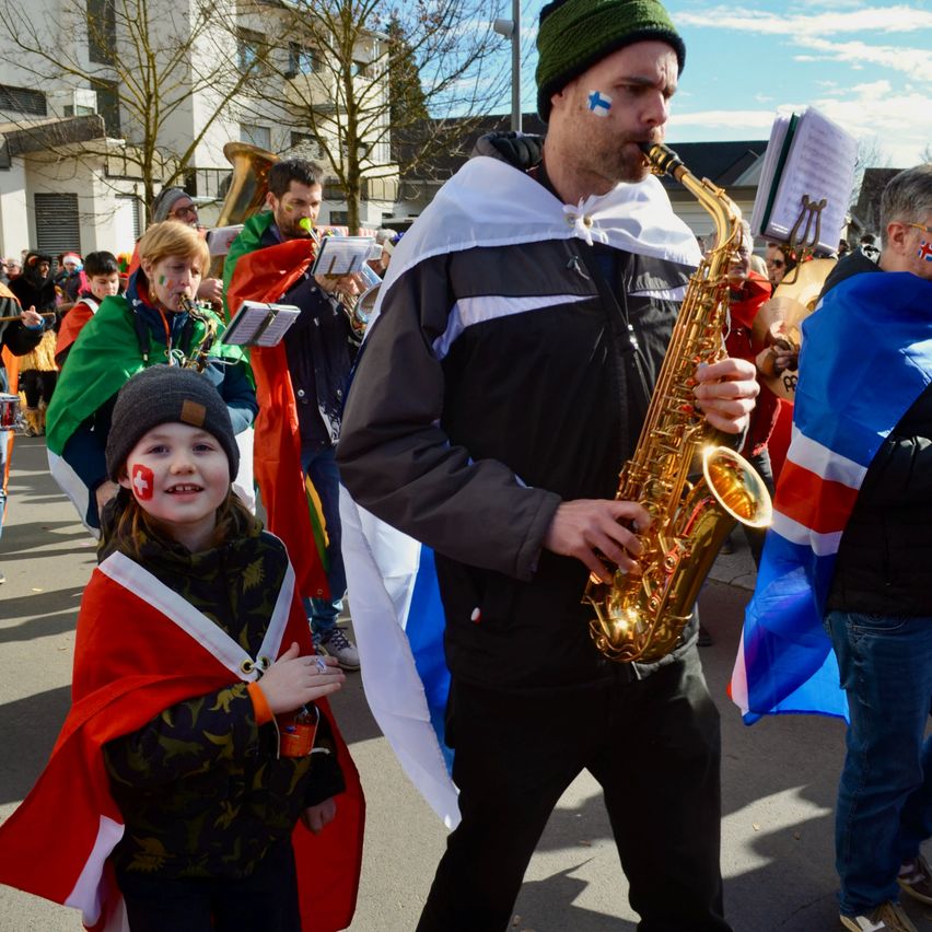 Ein Mann spielt Saxophon bei einer Parade, mit einer Menge im Hintergrund. Ein Kind mit einem Schweizer Flaggen-Cape steht neben ihm.
