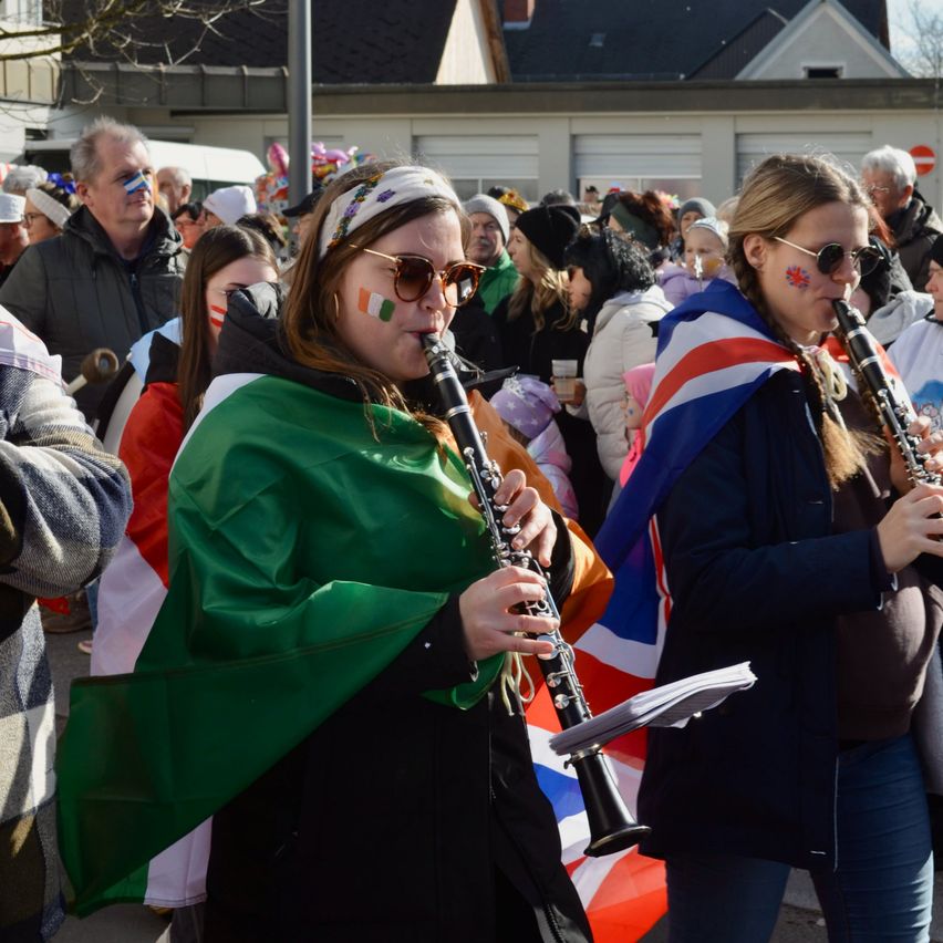 Eine Gruppe von Menschen, die in grünen und weißen Kostümen gekleidet sind, möglicherweise für einen Umzug, spielen Musikinstrumente, darunter eine Klarinette, während sie auf der Straße stehen.