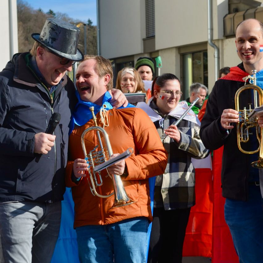 Mehrere Menschen in festlicher Kleidung, darunter ein Mann mit einer Trompete, stehen vor einem Gebäude. Sie lächeln und scheinen an einer Feier teilzunehmen.