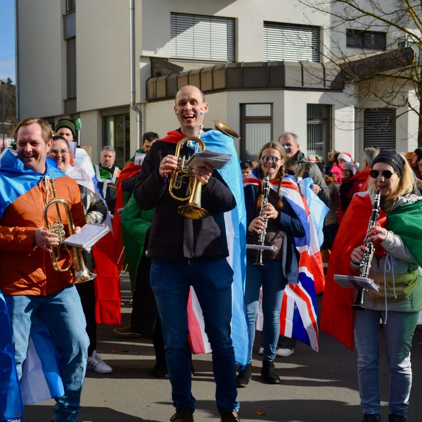 Eine Gruppe von Menschen in verschiedenen Kostümen, die Musikinstrumente halten, geht bei einem Umzug. Hinter ihnen ist ein Gebäude mit vielen Fenstern und einem Baum zu sehen.