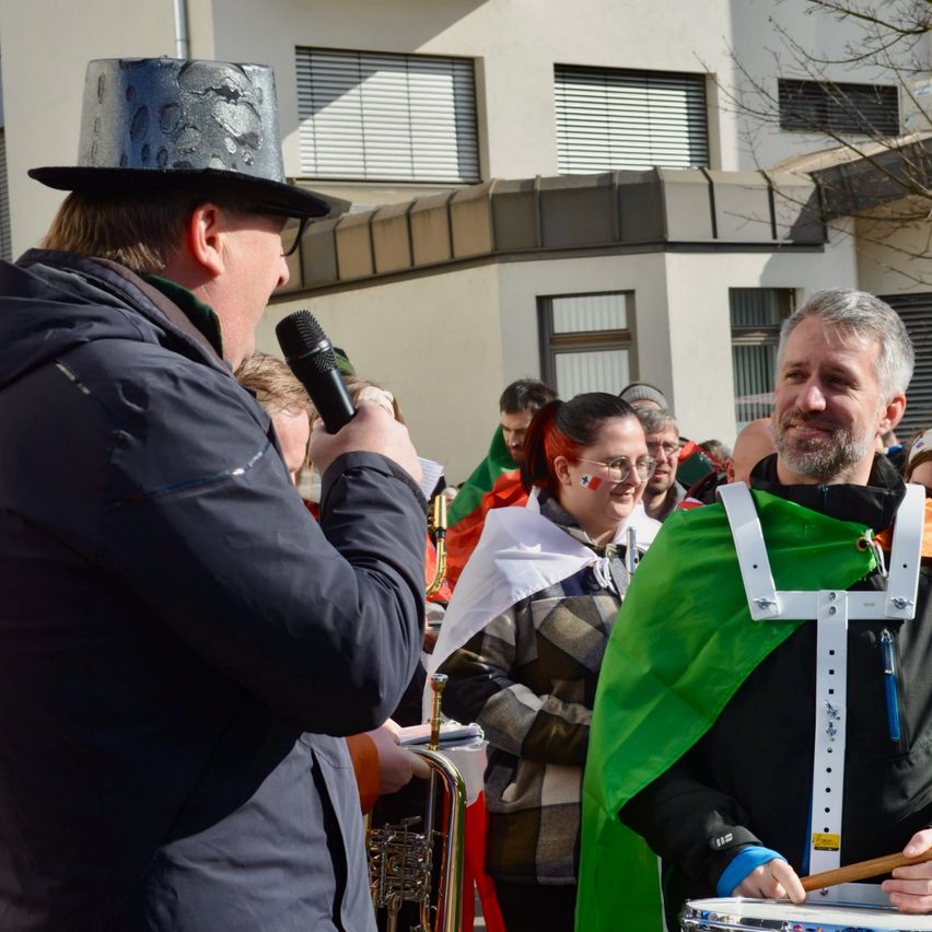 Ein Mann mit Mikrofon steht neben einem Schlagzeuger, der einen grünen Umhang trägt. Dahinter steht eine Frau mit Brille und einem roten Bandana im Haar, die zusieht. Im Hintergrund ein Gebäude mit Glasfenstern und einem Baum.