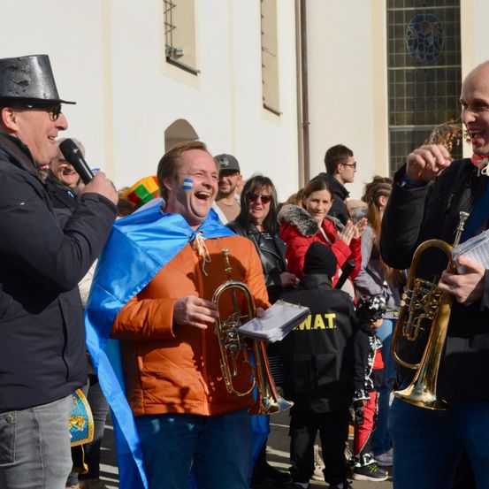 Eine Gruppe von Menschen steht im Freien; ein Mann hält eine Trompete, während ein anderer sie spielt, und ein dritter Mann mit einem Mikrofon spricht.
