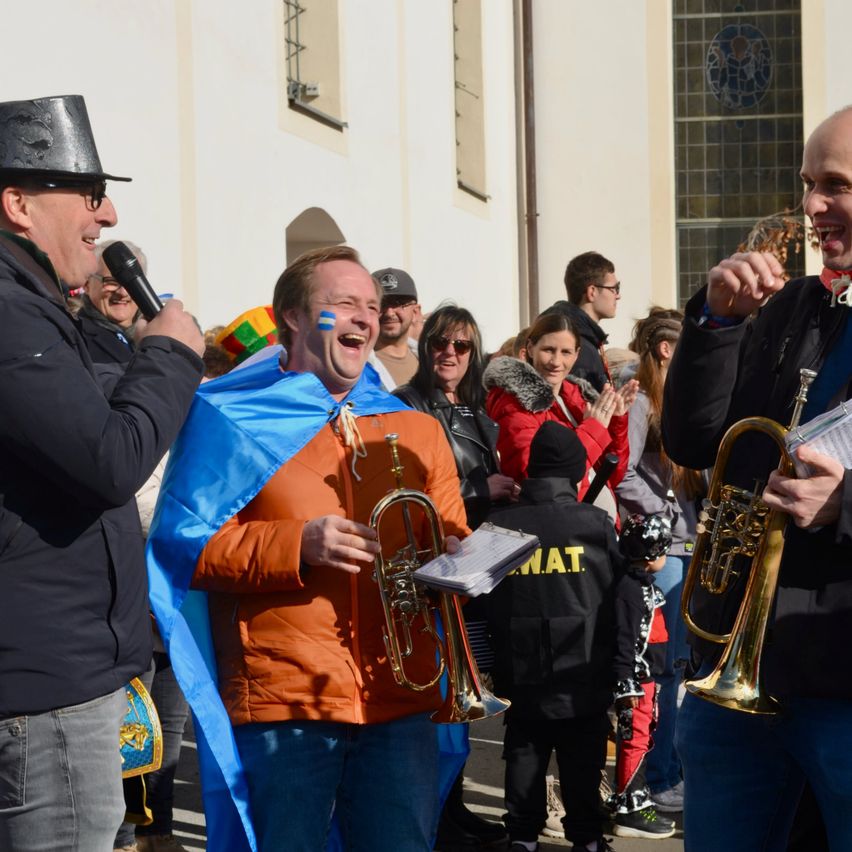 Eine Gruppe von Menschen steht im Freien; ein Mann hält eine Trompete, während ein anderer sie spielt, und ein dritter Mann mit einem Mikrofon spricht.