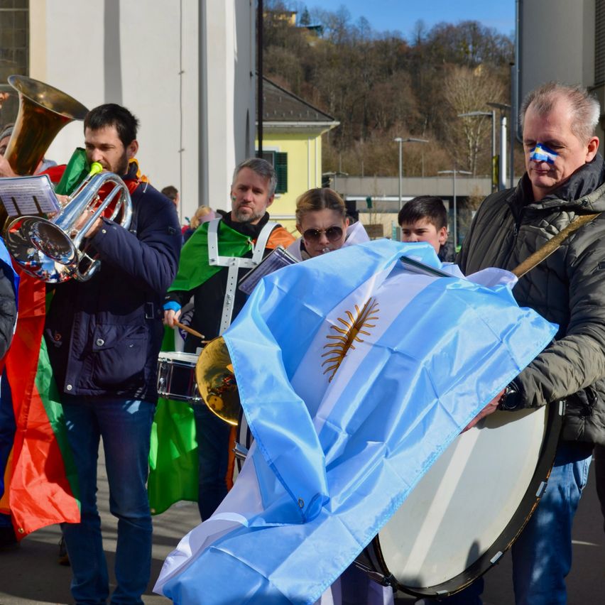 Eine Gruppe von Menschen ist im Freien, einige spielen Musikinstrumente. Ein Mann hält eine blaue Flagge mit Sonnensymbol. Ein anderer Mann hat blaue Farbe im Gesicht. Sie sind alle in Winterkleidung gekleidet.