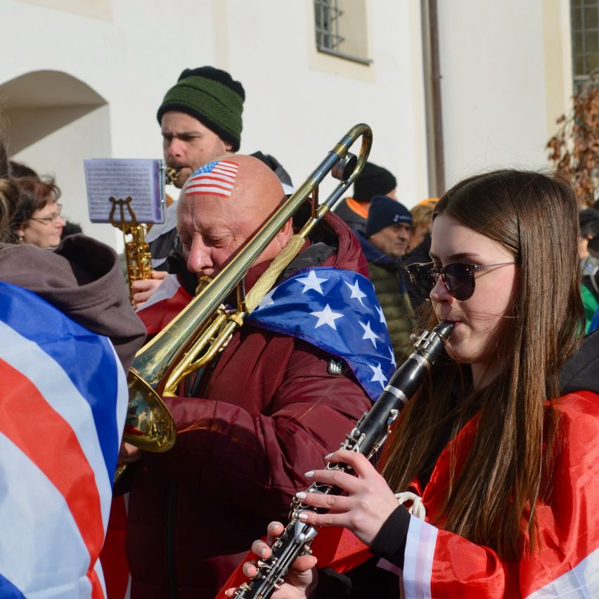 Eine Gruppe von Musikern spielt bei einer öffentlichen Veranstaltung, wobei ein kahlköpfiger Mann Posaune und ein Mädchen Klarinette spielt, beide tragen amerikanische Flaggen.