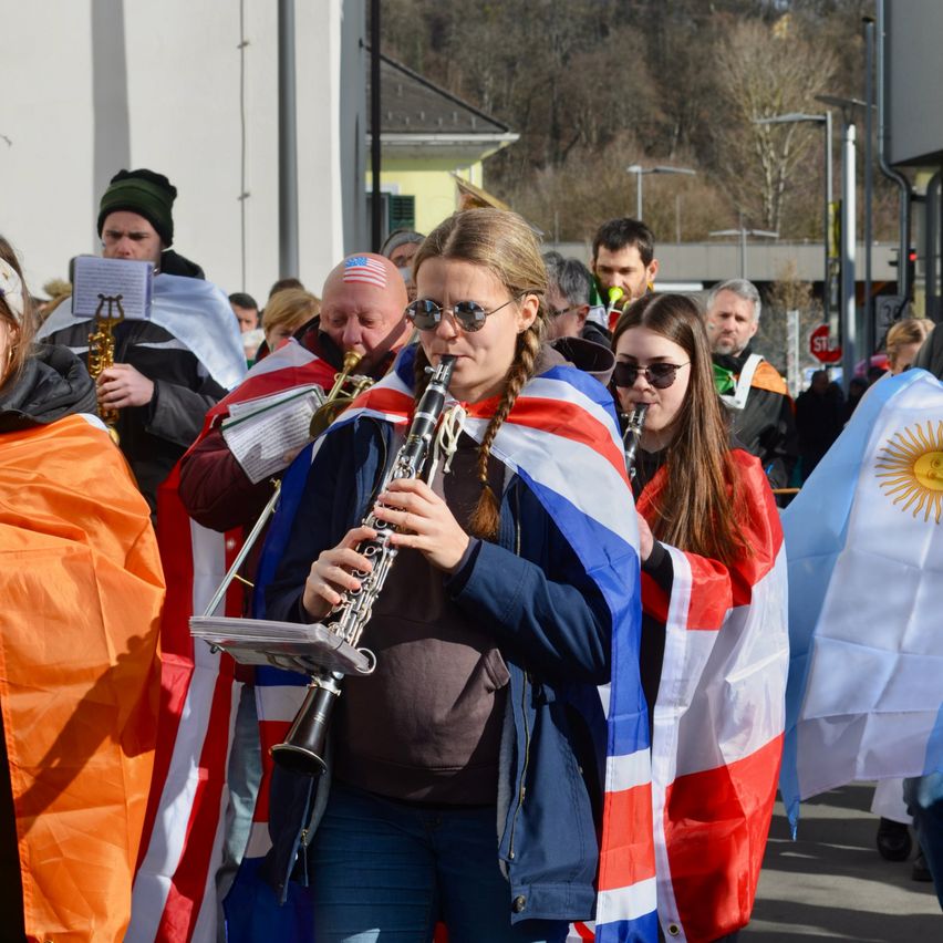 Eine Gruppe von Musikern marschiert bei einer Parade. Die Frau vorne spielt Klarinette, während andere Trompeten und Posaunen spielen. Sie sind in roten, weißen und blauen Kostümen gekleidet. Im Hintergrund befindet sich ein Gebäude und ein Stoppschild.