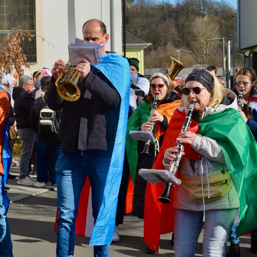 Eine Gruppe von Menschen in bunten Kostümen spielt Musikinstrumente bei einer Parade, mit einem Gebäude und Bäumen im Hintergrund.