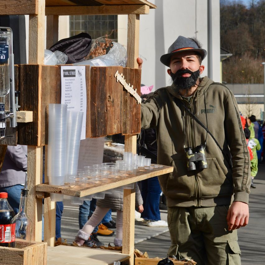 Ein Mann mit Hut und Jacke steht bei einem Holzstand mit Bechern und einer Flasche. Hinter ihm laufen Menschen auf einer Straße. Bäume und Gebäude sind im Hintergrund.