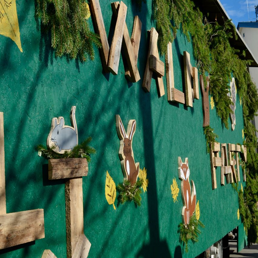 Eine grüne Wand mit Holzbuchstaben, die 'WALLY' buchstabieren. Darunter steht ein hölzerner Pfosten mit einer Hasenfigur. Daneben befindet sich ein hölzerner Fuchs. Grünes Laub und Blätter schmücken die Wand.