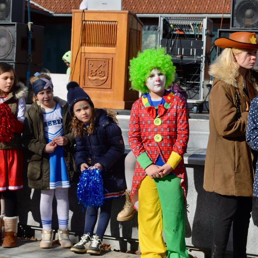 Eine Gruppe von Kindern und ein Clown stehen zusammen, vielleicht für ein Foto. Dahinter steht ein hölzernes Podium mit einem Symbol und einigen Lautsprechern.