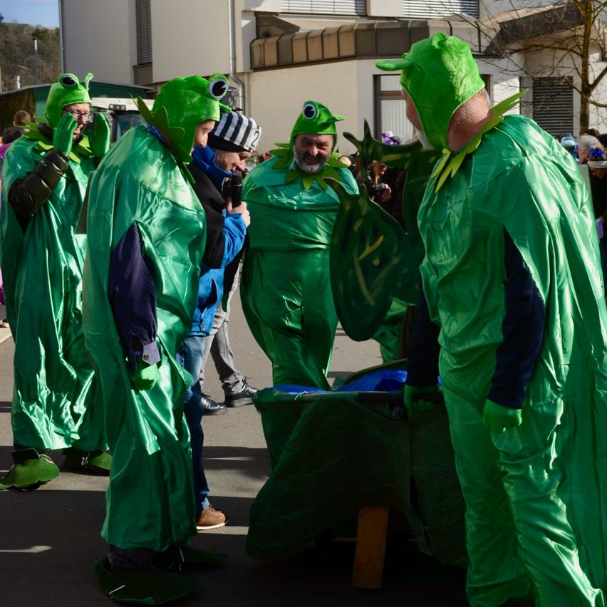 Eine Gruppe von Menschen in grünen Froschkostümen versammelt sich im Freien, möglicherweise für eine Parade oder Veranstaltung. Ein Mann macht ein Foto mit einer Kamera.
