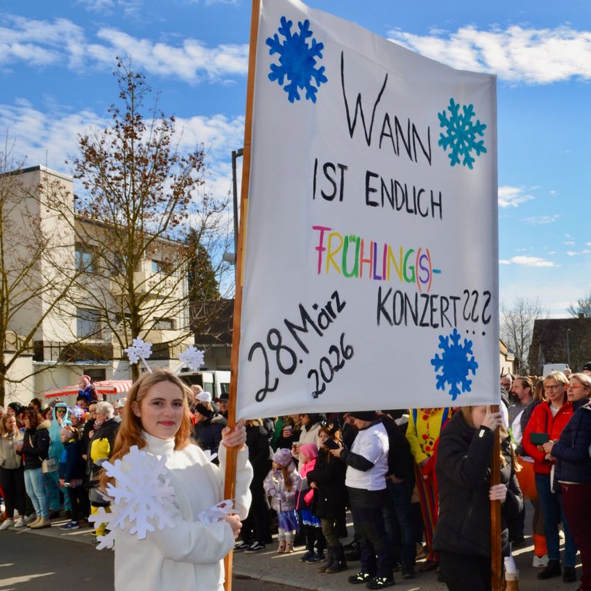 Eine Frau in einem weißen Winteroutfit hält ein Banner mit Schneeflocken und dem Text 'Wann ist endlich Frühlings-Konzert 2026?' vor einer Menschenmenge, mit Gebäuden und einem blauen Himmel im Hintergrund.