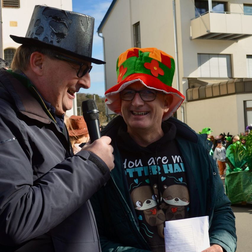 Zwei lächelnde Männer, einer hält ein Mikrofon und Papier, der andere trägt einen bunten Hut. Sie stehen vor einem Gebäude mit Menschen und Pflanzen im Hintergrund.