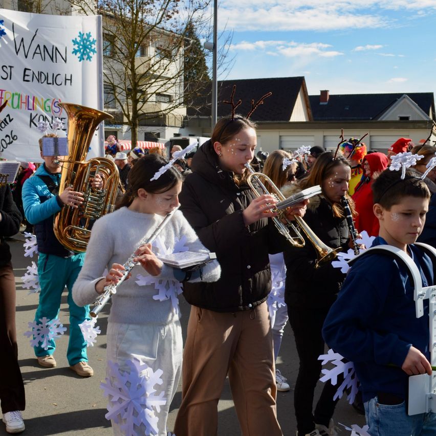 Eine Gruppe von Menschen in Kostümen, die bei einer Parade Musikinstrumente spielen. Ein Mädchen mit einer Schneeflockengirlande und einer Trompete, ein anderes mit einer Flöte und ein Junge mit einem Tuba. Ein Banner mit Schneeflocken und Text ist im Hintergrund.