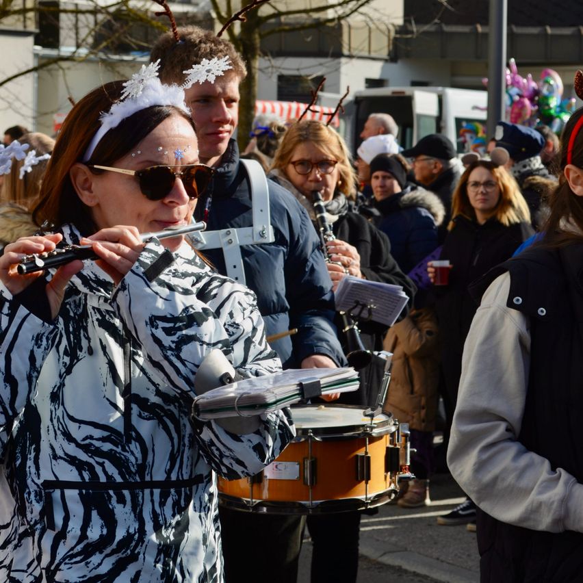 Eine Frau in einem Zebra-Jackett spielt Flöte, umgeben von einer Menschenmenge in Winterkleidung und Weihnachtskopfbändern, mit sichtbaren Musikinstrumenten.