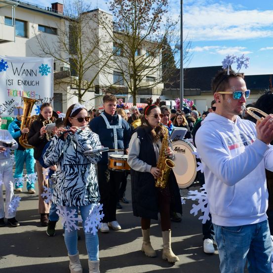 Eine Blaskapelle spielt auf einer Straße mit Gebäuden und Bäumen im Hintergrund. Die Teilnehmer sind in Winterkleidung gekleidet, einige mit Schneeflocken an ihren Jacken.