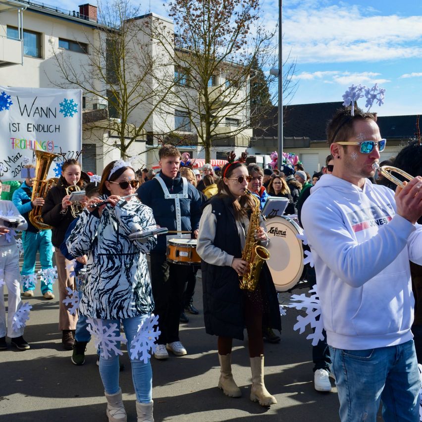 Eine Blaskapelle spielt auf einer Straße mit Gebäuden und Bäumen im Hintergrund. Die Teilnehmer sind in Winterkleidung gekleidet, einige mit Schneeflocken an ihren Jacken.