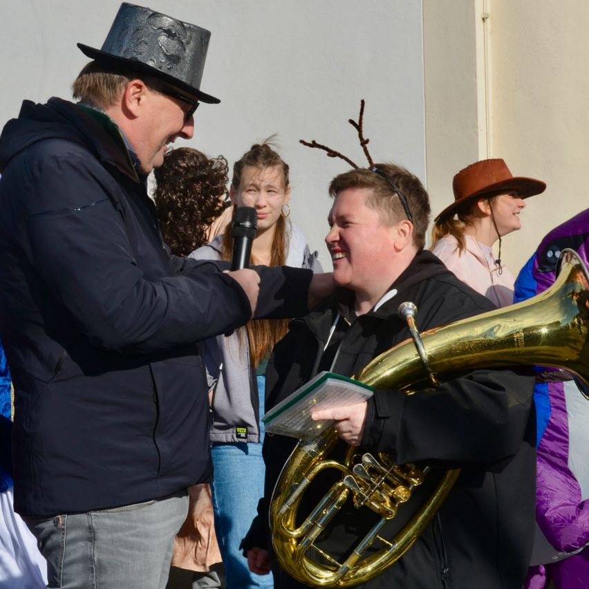 Ein Mann mit Hut und Mikrofon spricht mit einer lächelnden Frau, die ein Tuba hält, während andere zusehen.