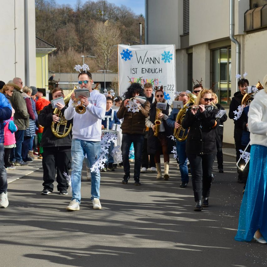 Eine Gruppe von Menschen in Winterkleidung geht spielend auf der Straße. Ein Schild dahinter lautet 'Wann ist endlich'.