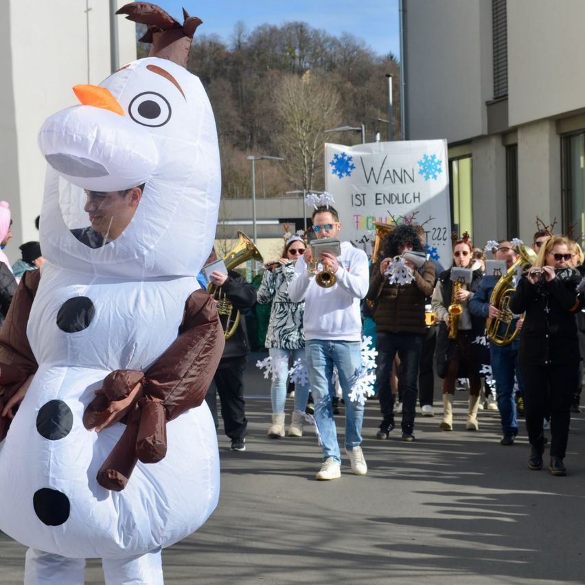 Eine Person in einem großen Olaf-Kostüm führt eine Marschkapelle an und spielt Instrumente wie Posaunen und Tuben bei einer festlichen Parade im Freien. Dahinter steht auf einem Banner 'Wann ist endlich'.