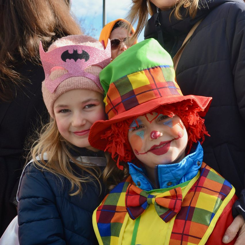 Ein junges Mädchen mit einer rosa Batman-Mütze und ein Junge in einem farbenfrohen Clownskostüm posieren zusammen bei einer Veranstaltung im Freien.