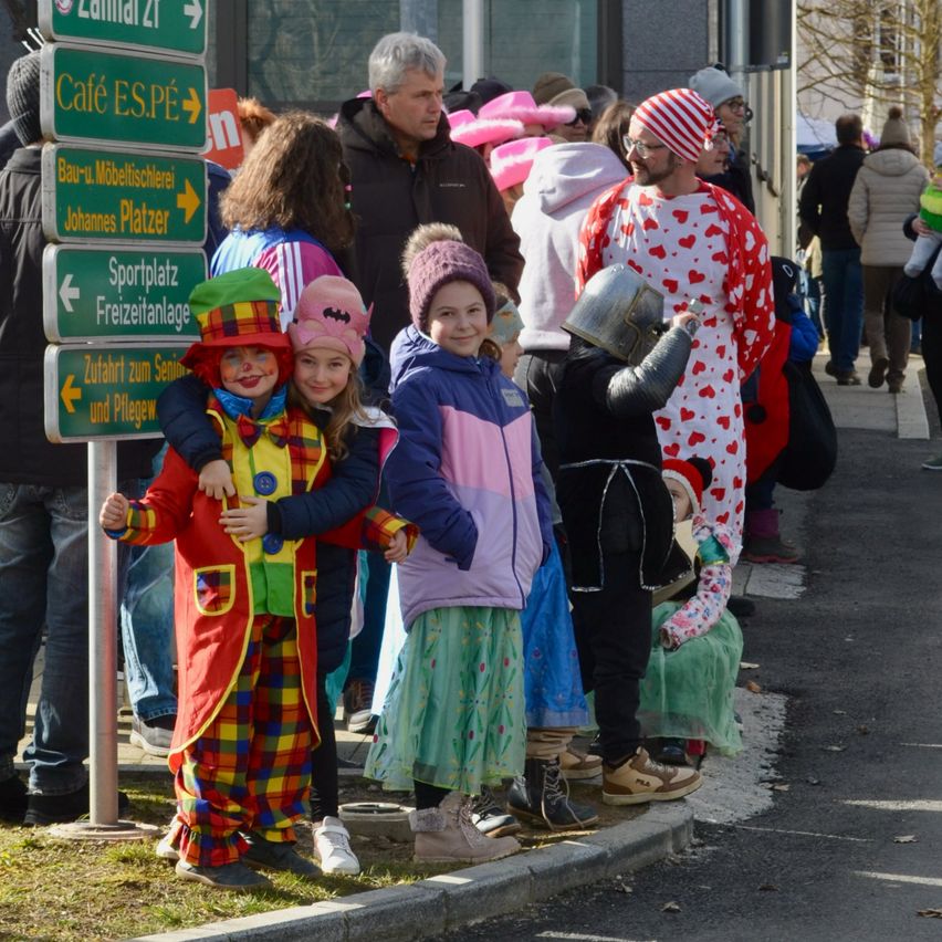 Eine Gruppe von Kindern und Erwachsenen in Kostümen steht auf einem Bürgersteig. Einige tragen Hüte und andere halten Requisiten. Hinter ihnen steht ein grünes Schild mit Pfeilen.