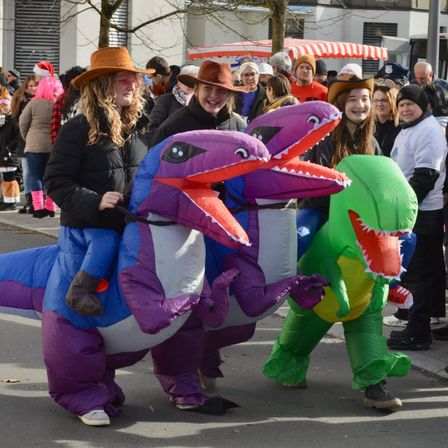 Menschen in Dinosaurierkostümen laufen in einer Parade. Zwei Frauen sitzen auf dem lila Dinosaurier, während eine andere Frau in einem grünen Dinosaurierkostüm steht. Dahinter schauen Leute von den Seiten zu.