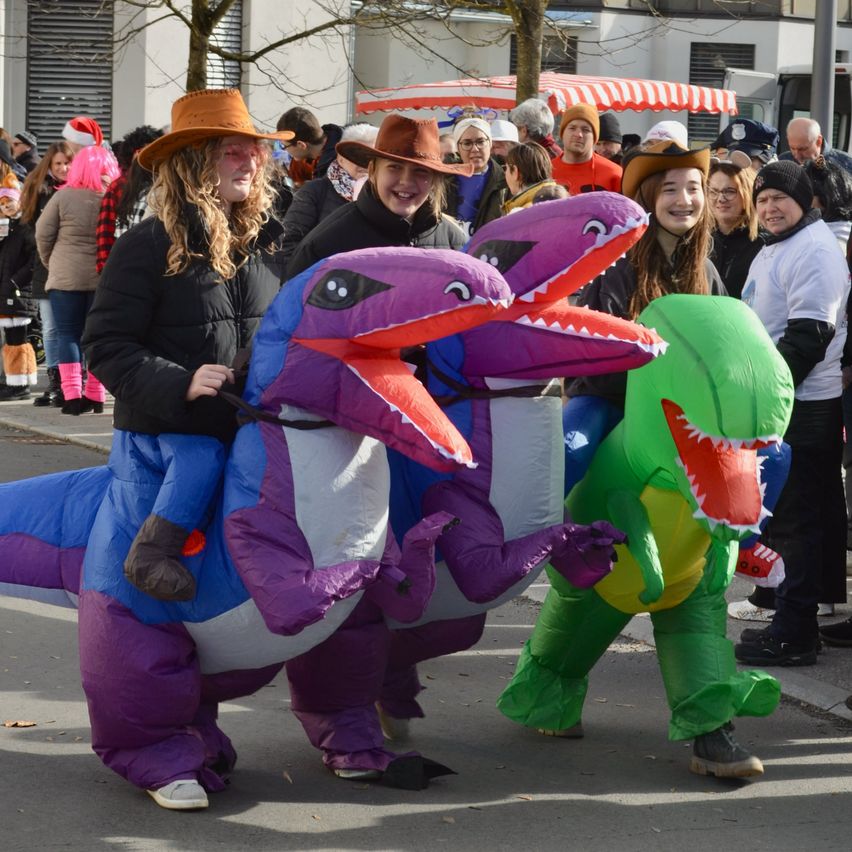 Menschen in Dinosaurierkostümen laufen in einer Parade. Zwei Frauen sitzen auf dem lila Dinosaurier, während eine andere Frau in einem grünen Dinosaurierkostüm steht. Dahinter schauen Leute von den Seiten zu.