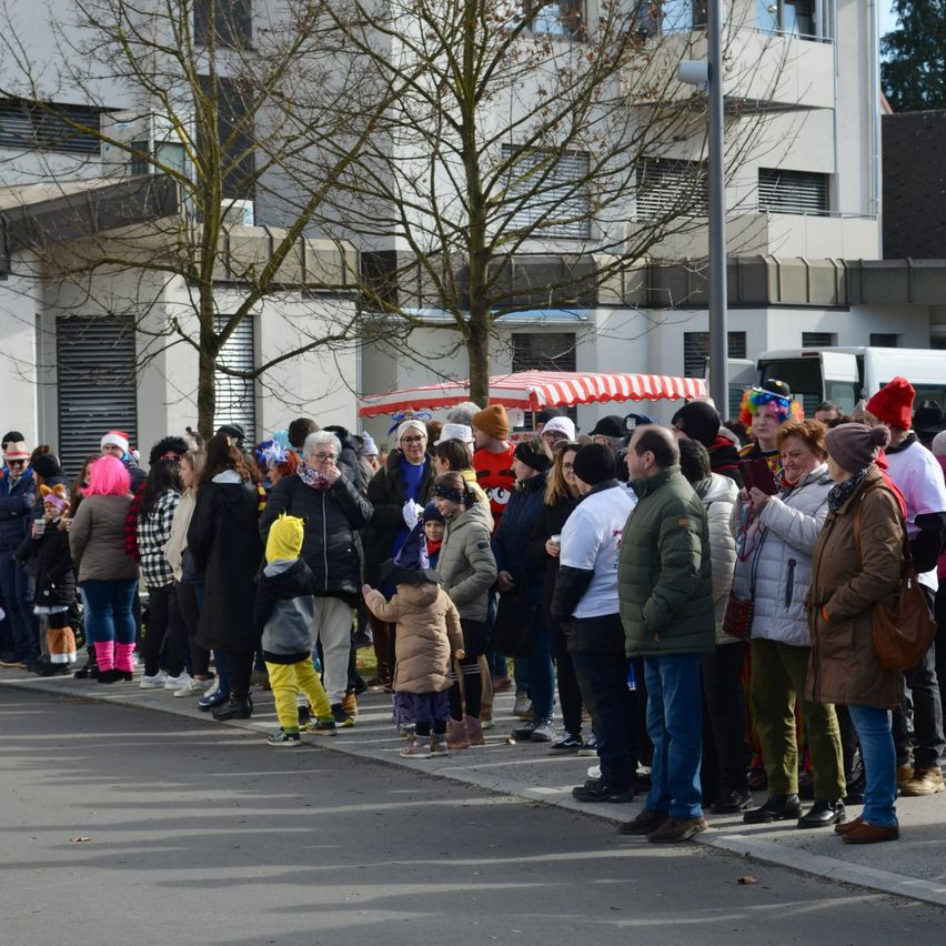 Eine Menschenmenge, einschließlich Kinder und Erwachsene, steht in einer Reihe auf dem Bürgersteig. Sie tragen alle Winterkleidung, Mützen und Schuhe. Das Gebäude hinter ihnen hat Glasfenster und ein rot-weißes Zelt. Das Gebiet ist von Bäumen umgeben.